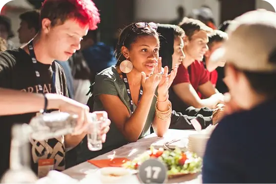 A group of people sitting at a table, talking and pouring drinks. A woman gestures while listening. A salad and table number 17 are visible.