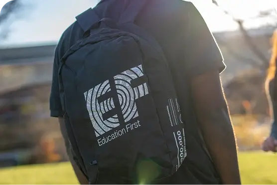 Person wearing a black backpack with a white "Education First" logo, standing outside on a sunny day.
