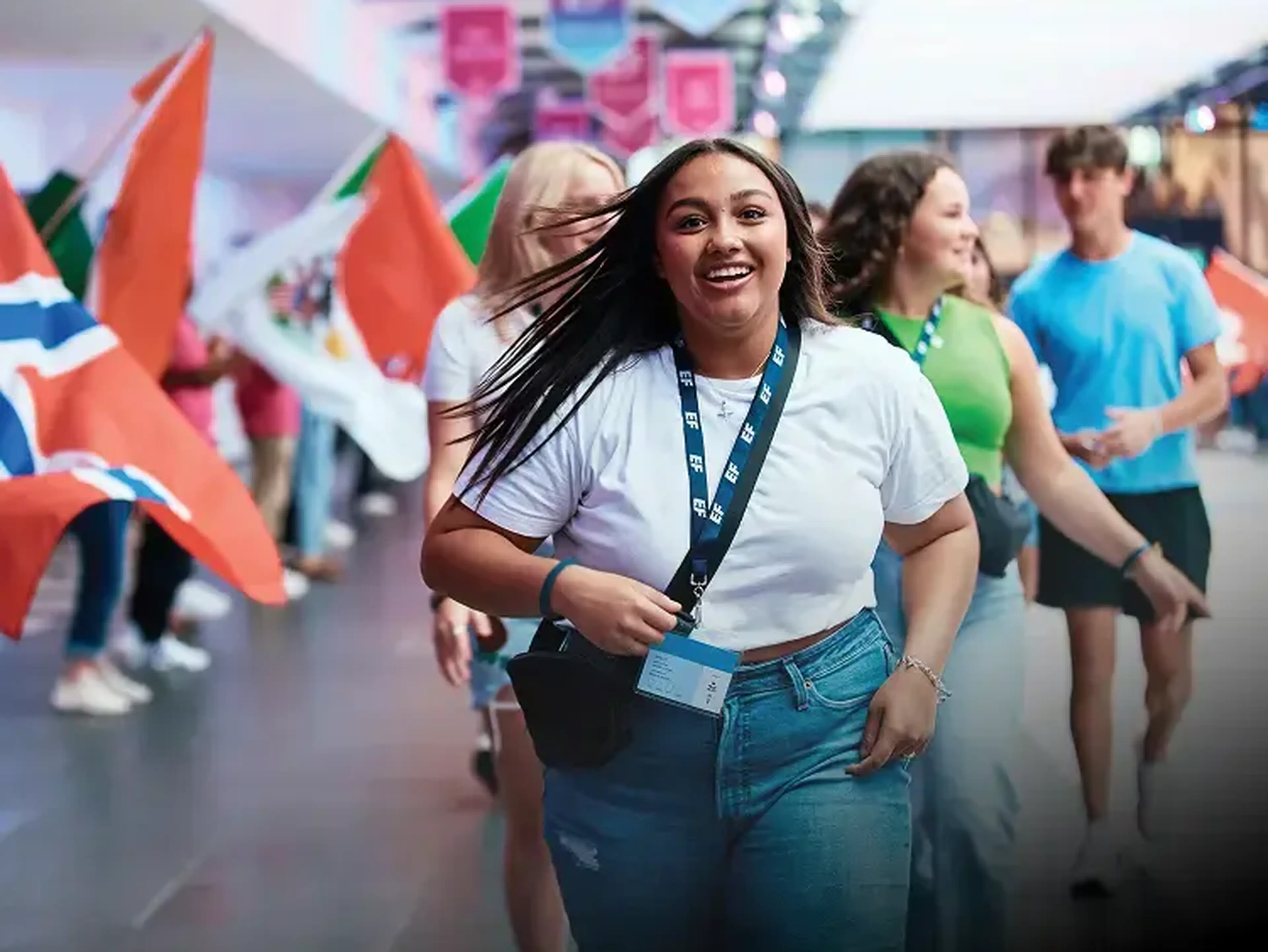 A smiling woman in a white shirt and jeans walks indoors, surrounded by people holding flags.