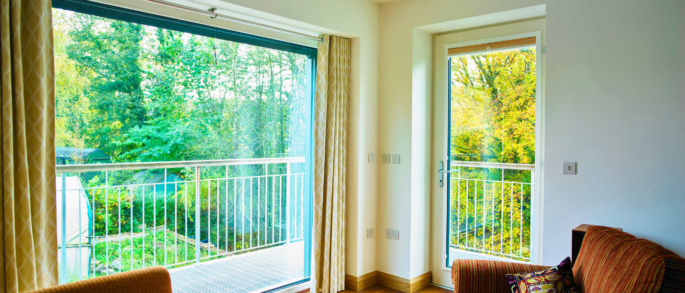 Bright room with large windows and a door leading to a balcony with a green, leafy view. A brown sofa is partially visible on the right.