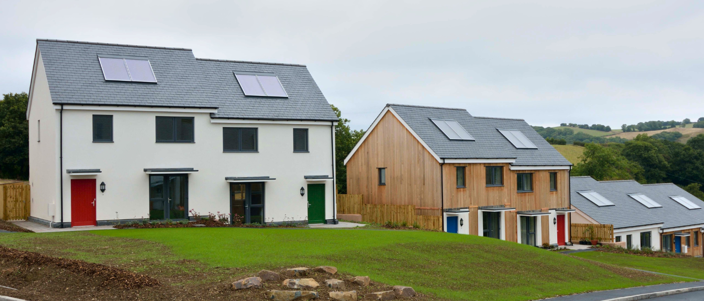 Modern houses with solar panels on sloped roofs, featuring green lawns and surrounding trees under a cloudy sky.
