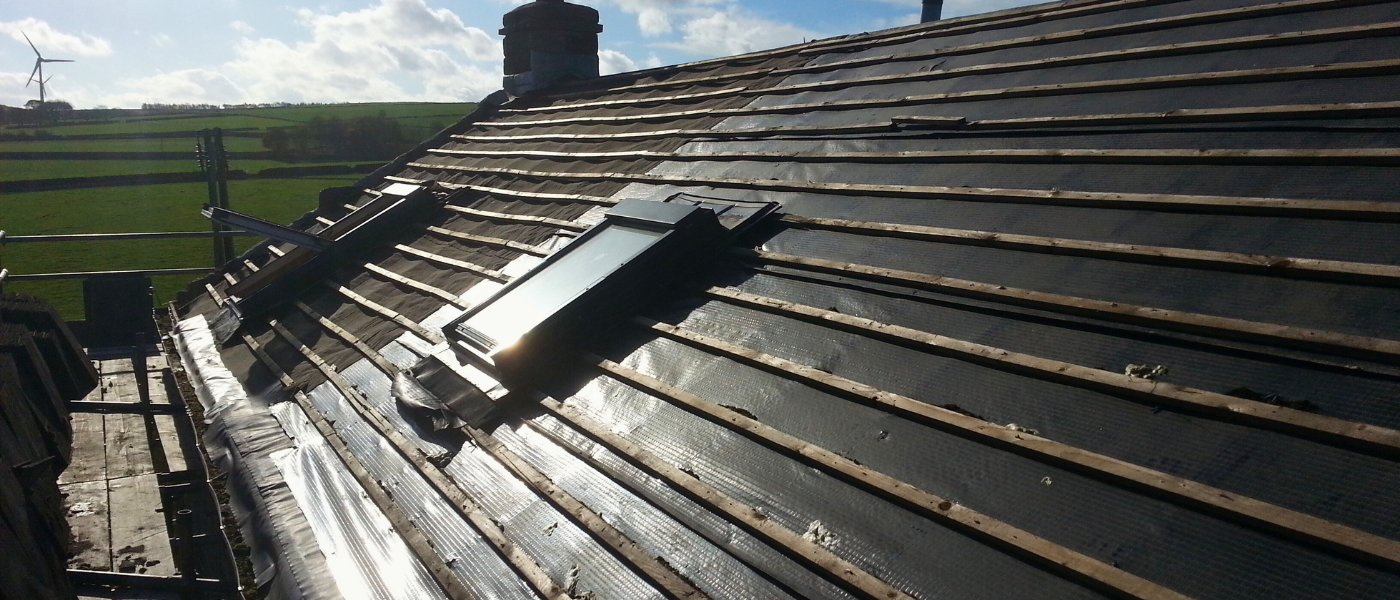 A sloped roof under construction with exposed wooden battens and installed skylights, set against a backdrop of fields and a wind turbine.