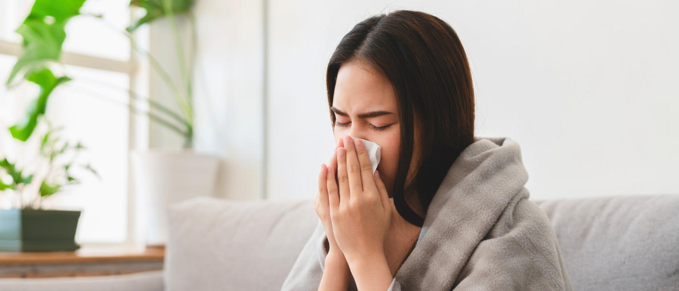 A woman wrapped in a blanket sits on a couch, sneezing into a tissue. A potted plant is visible in the background.