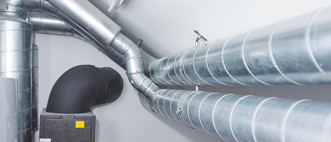 Metallic ventilation ducts and a black insulated pipe are installed against a white ceiling, with fittings and clamps visible.