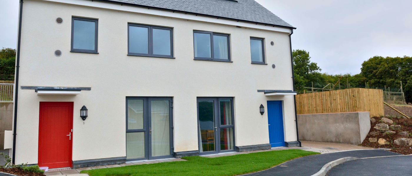 Modern duplex with white facade, featuring red and blue front doors, large windows, and a small lawn, set against a cloudy sky.
