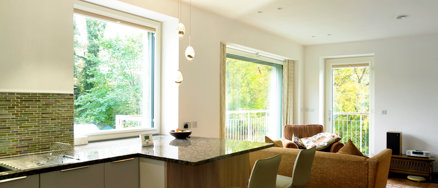 Modern living room with large windows, a cozy sofa, pendant lights, and a kitchen island. Outside, lush greenery is visible.