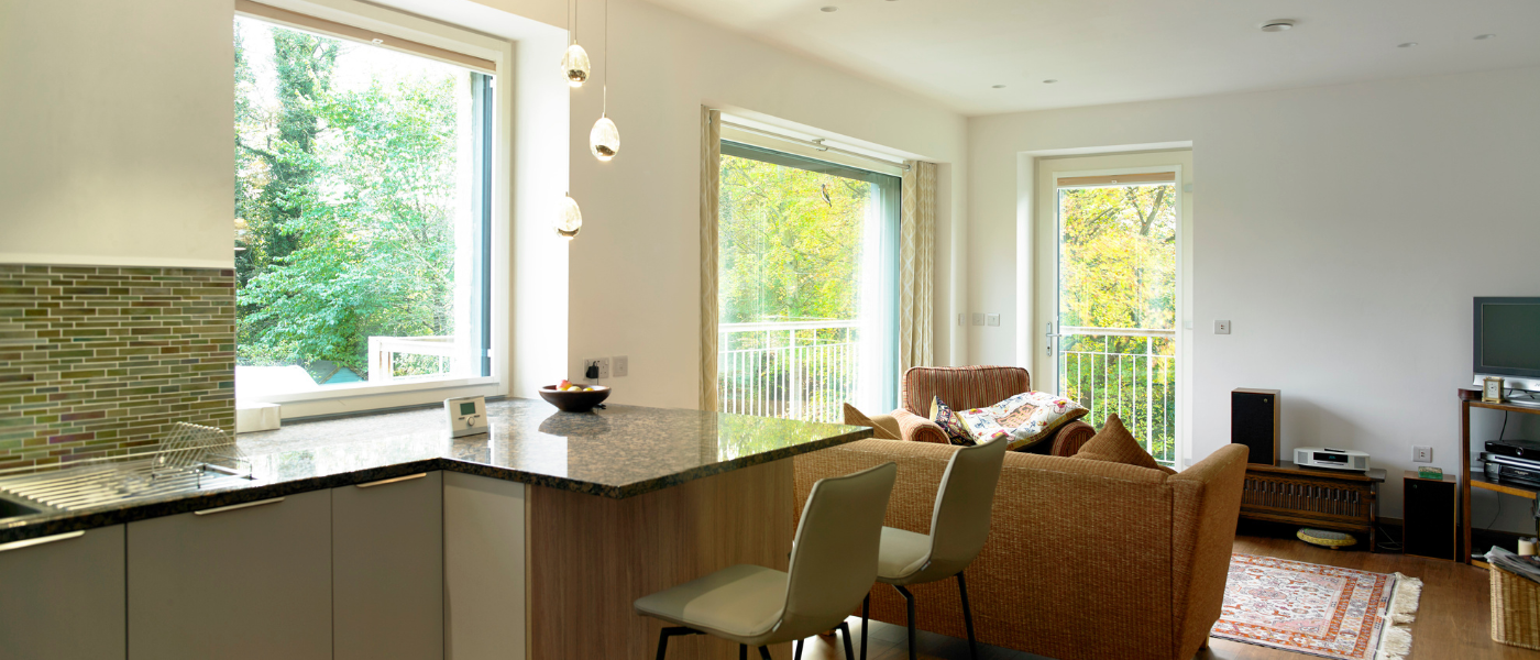 Modern open-plan living room and kitchen with large windows, brown sofa, dining area, and lush greenery visible outside.