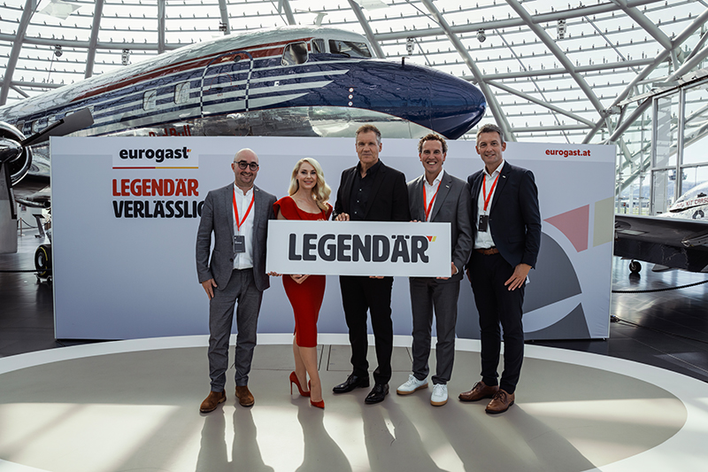 Fünf Personen halten ein Schild mit der Aufschrift "LEGENDÄR" vor einem Flugzeug.