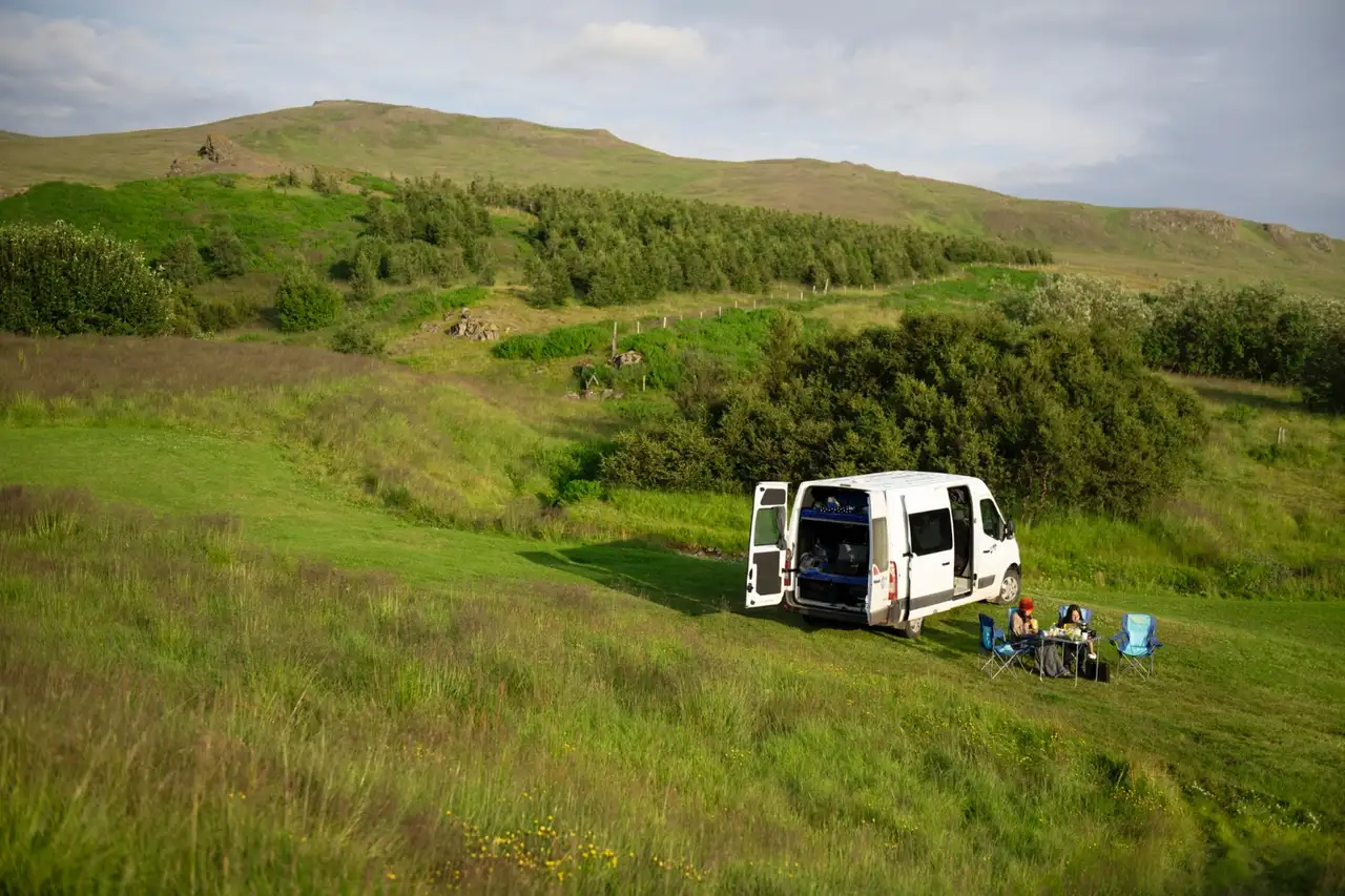 Ein Camper steht in der Natur und zwei Personen sitzen in Campingstühlen daneben