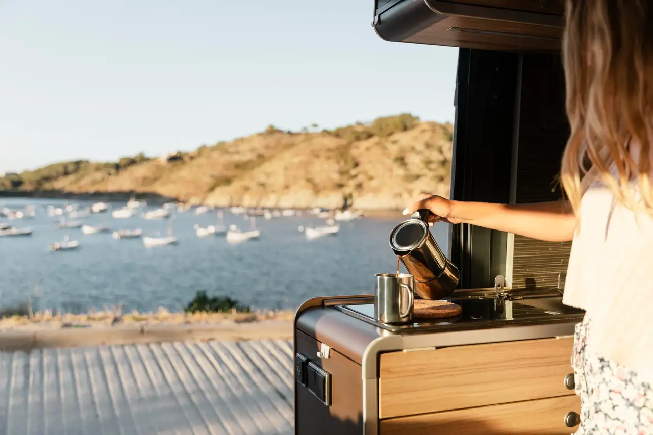 Person gießt sich einen Kaffe in ihrem Van ein und schaut dabei auf das Meer, auf dem sich viele weiße Boote befinden