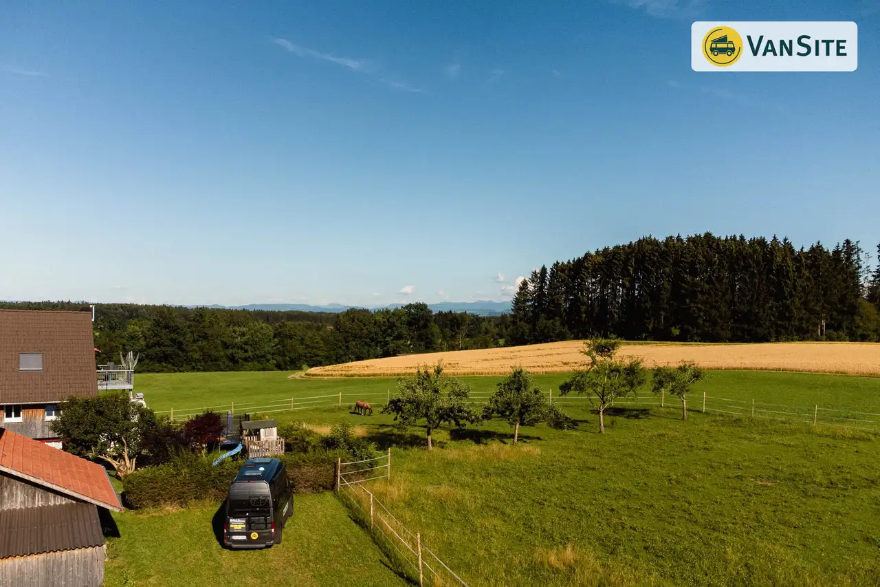 Ein Camper steht auf einem Feld, links Häuser, dahinter Pferde