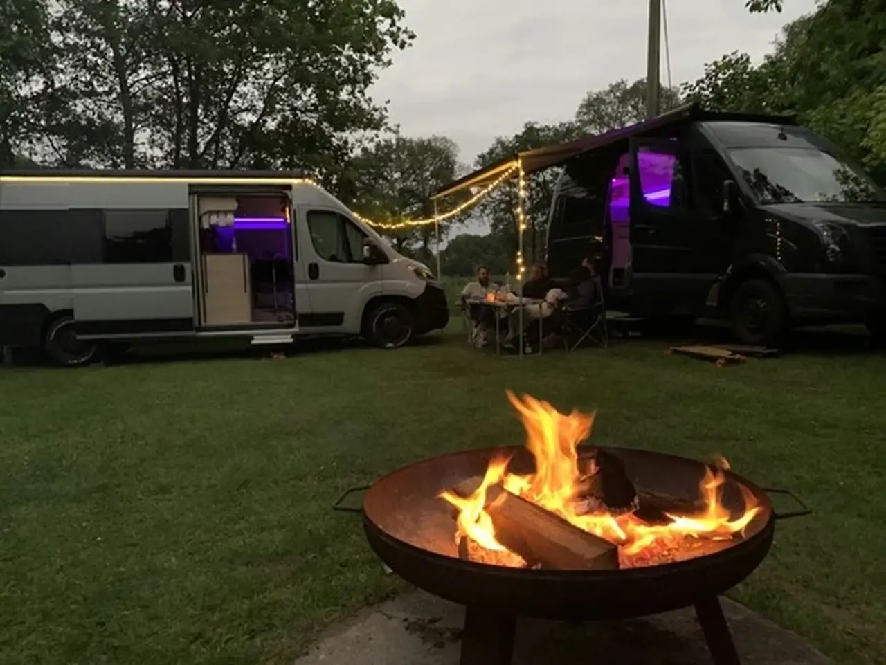 Zwei Camper stehen auf einer Wiese, eine Lichterkette ist zwischen ihnen aufgespannt und ein Lagerfeuer brennt im Vordergrund
