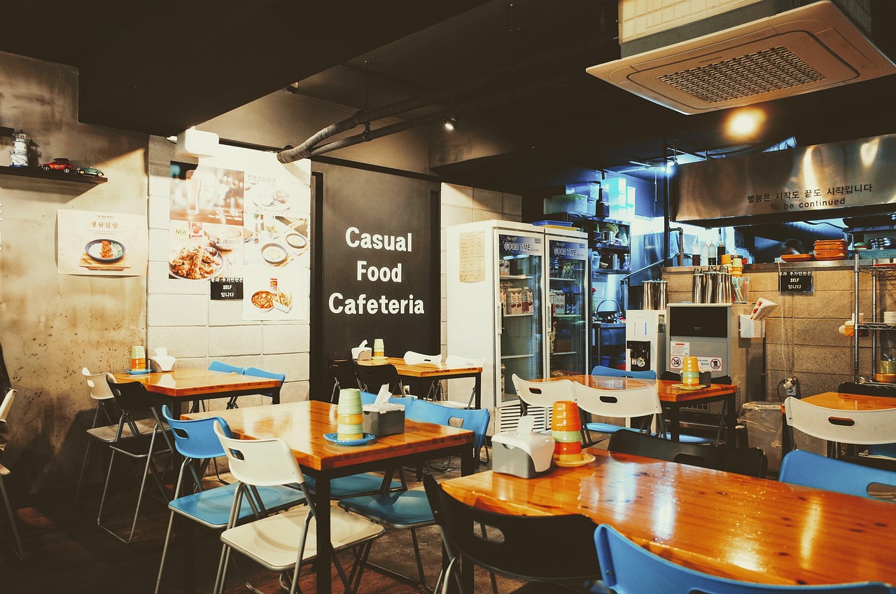 Casual cafeteria with wooden tables, colourful chairs, and kitchen area. Walls display food images and text. Cosy, inviting atmosphere.