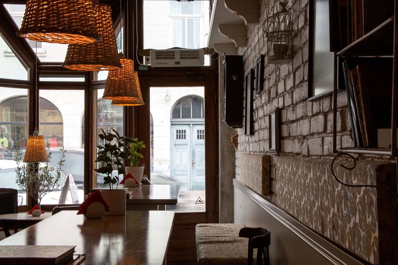 Cosy restaurant interior with wicker lamps, wooden tables, brick walls, and large windows overlooking a street.