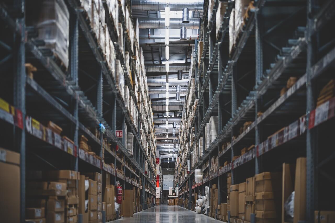 A large warehouse aisle with tall shelves filled with various boxes and packages, extending into the distance under bright overhead lighting.