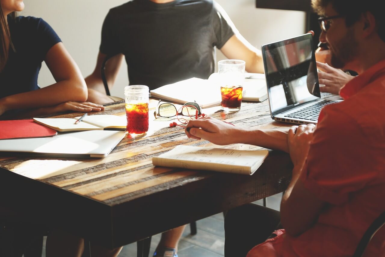 People sitting around a wooden table with notebooks, a laptop, and drinks, engaged in a discussion.
