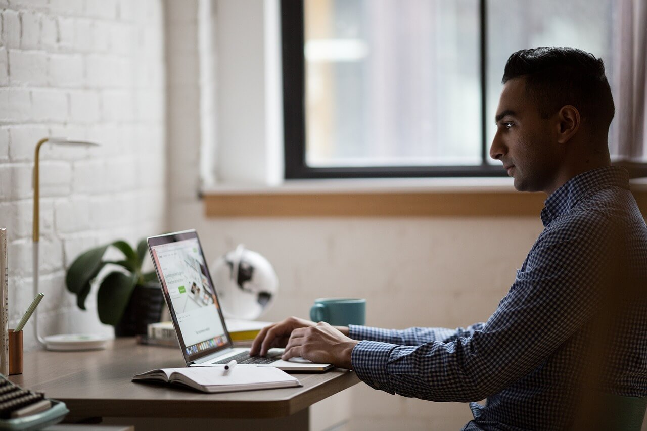 Man working on a laptop at a desk with a notebook and mug, in a bright room with a window.