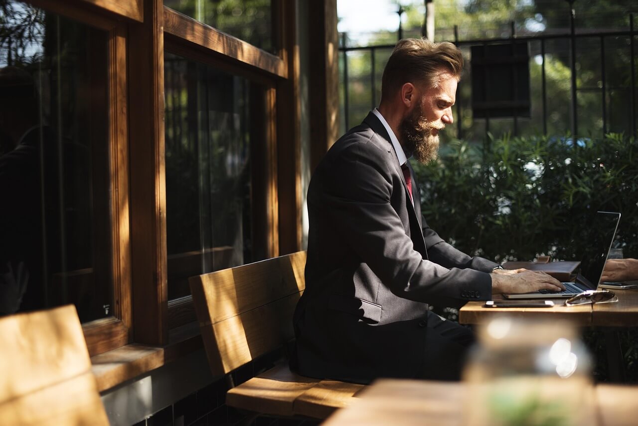 Man in a suit working on a laptop at an outdoor cafe, with sunlight streaming through the windows and greenery in the background.