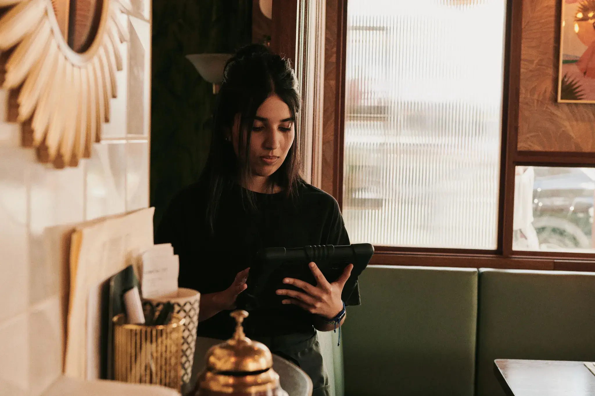 Woman in a cafe holding a tablet, standing near a window with soft lighting and decorative elements in the background.