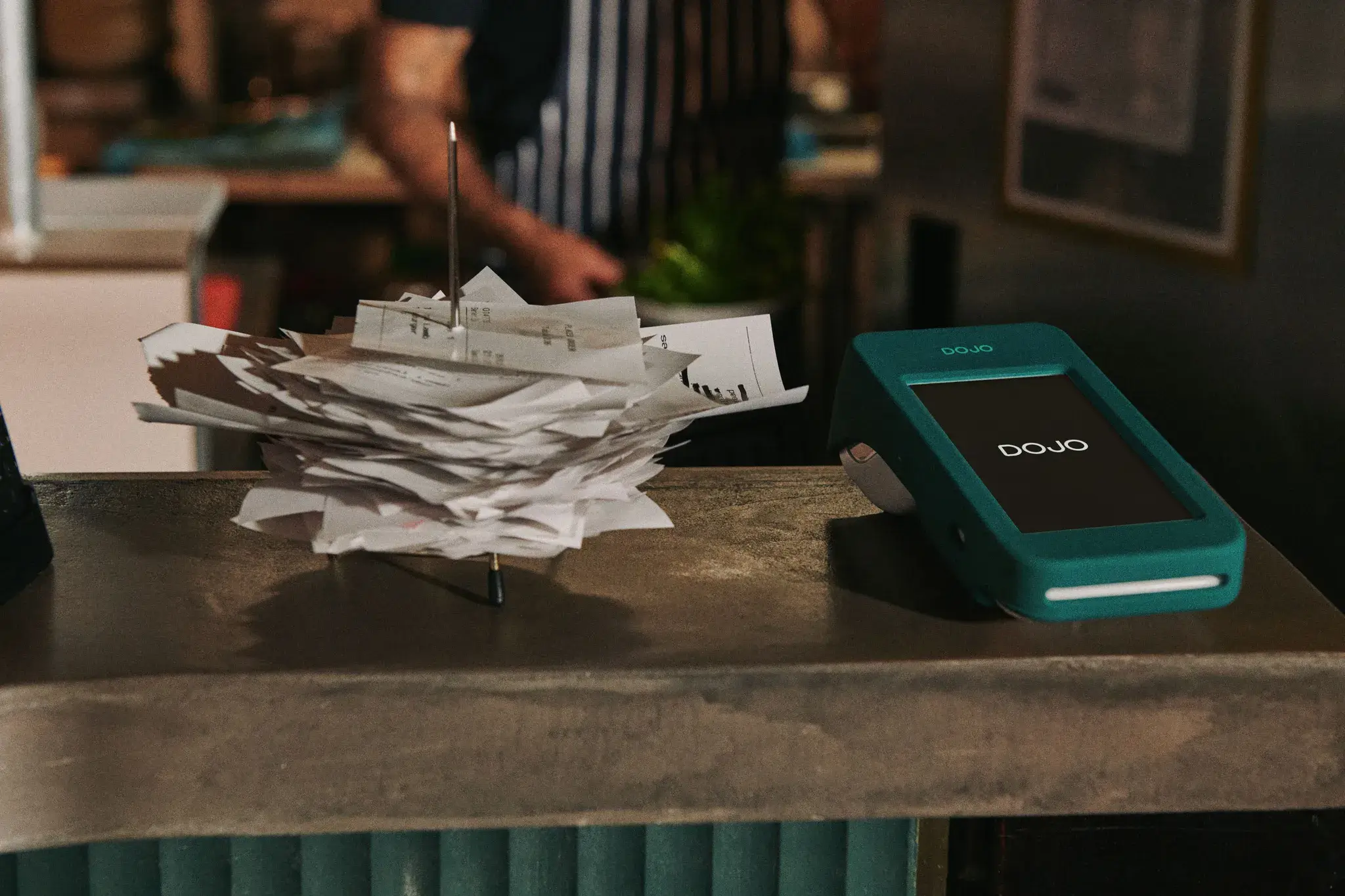 A stack of paper receipts on a spike next to a teal Dojo card machine on a counter.