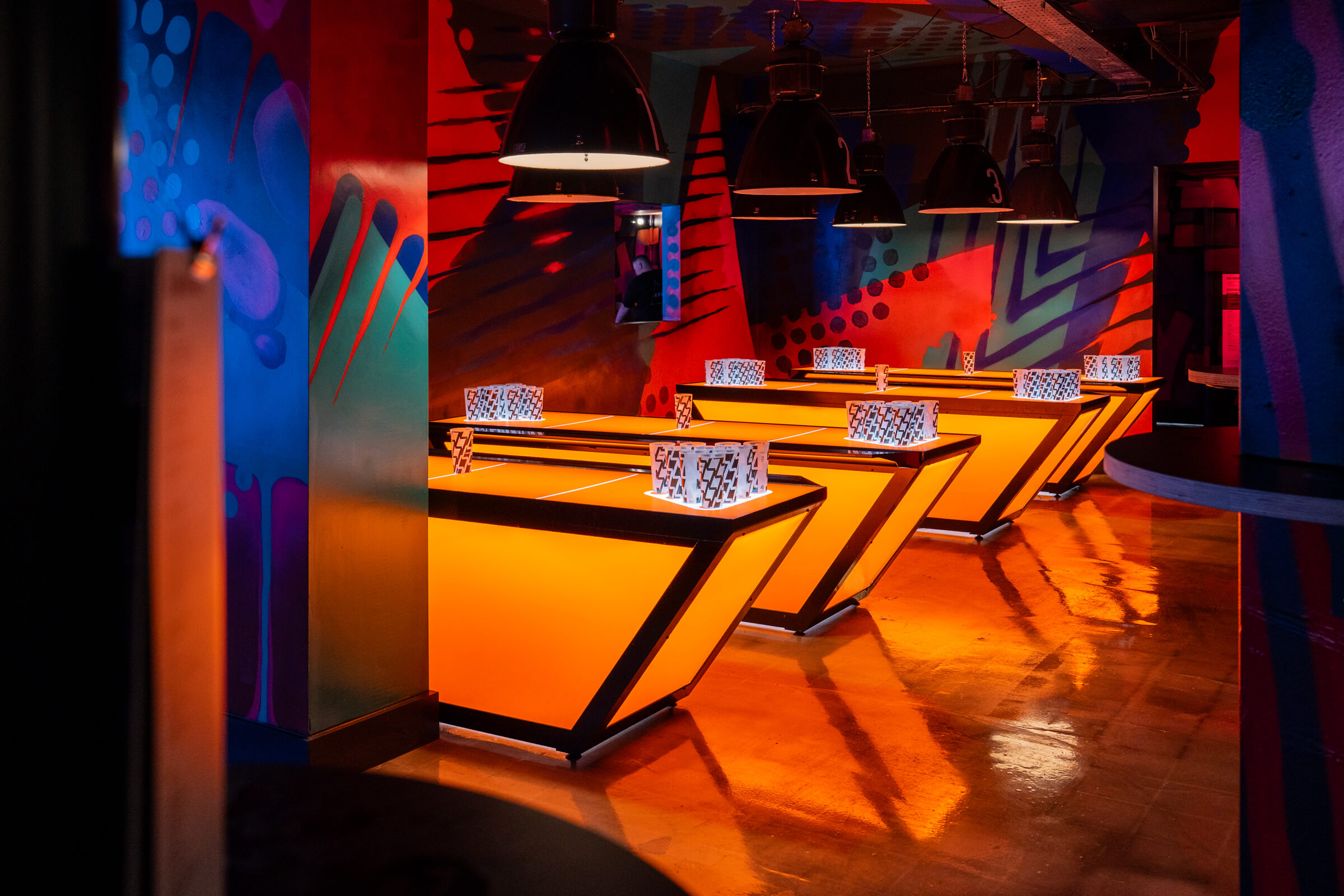 Vibrant bar interior with illuminated orange tables, colourful wall art, and black pendant lights creating a lively atmosphere.