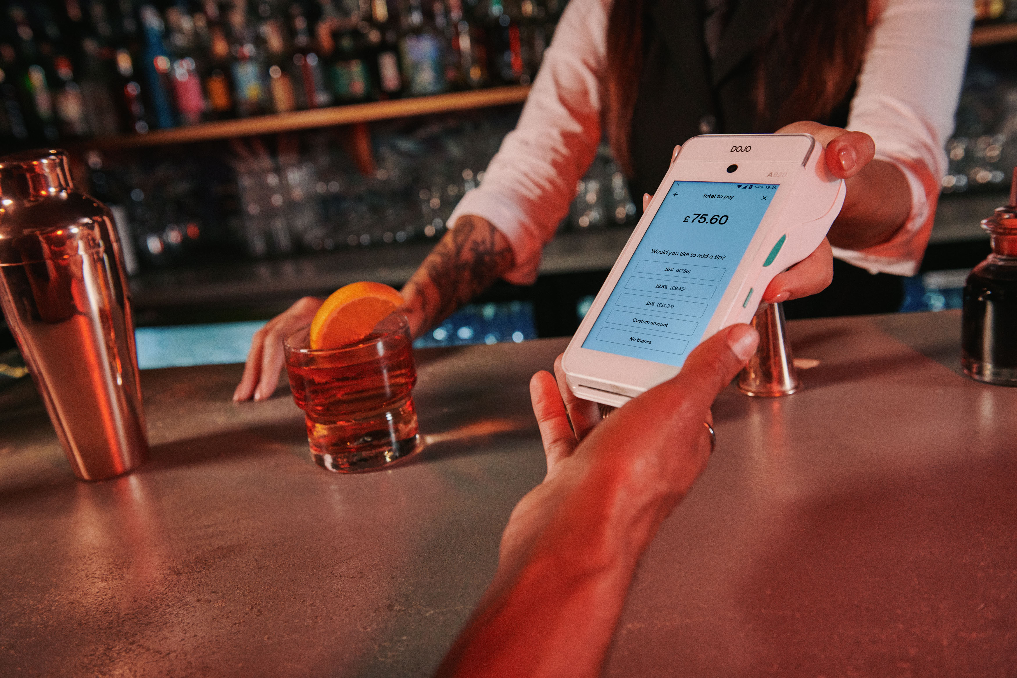 Bartender handing a Dojo card reader to a customer at a bar counter, with a cocktail garnished with an orange slice nearby.