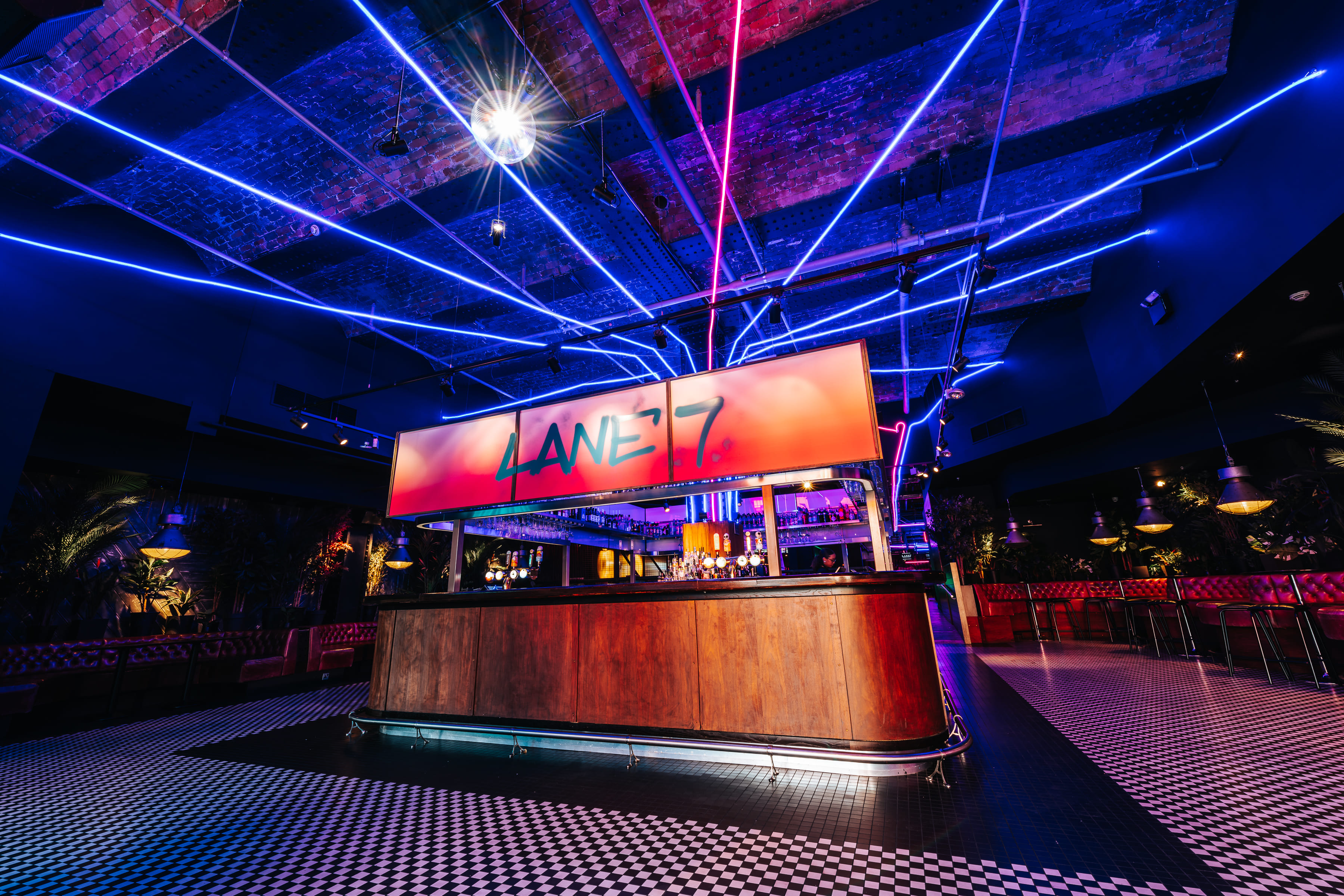 A vibrant bowling alley interior with neon lights, a checkered floor, and a bar area under a glowing "LANE 7" sign.