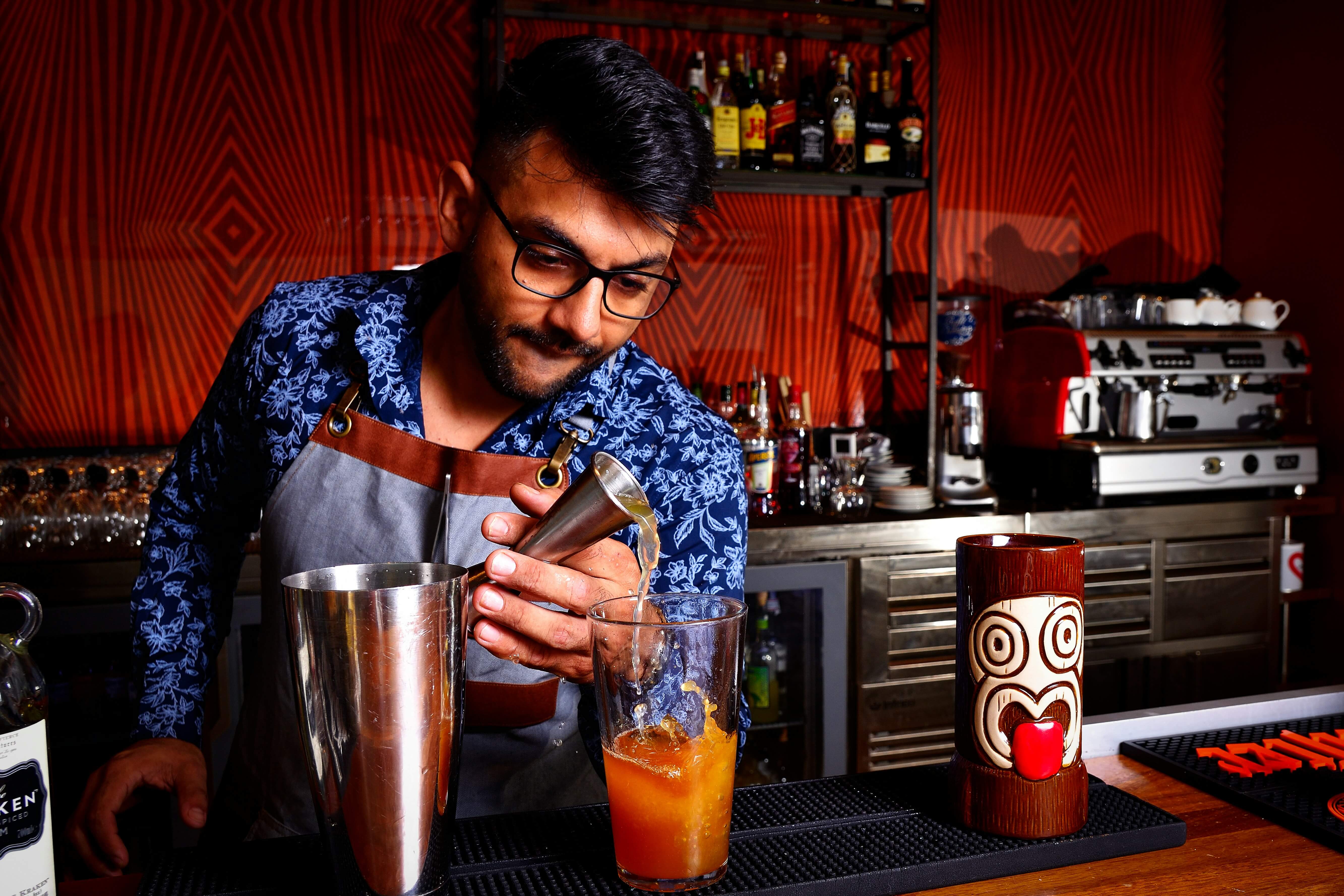 Bartender with glasses and a blue patterned shirt pours a drink into a glass at a bar, surrounded by bottles and bar equipment.