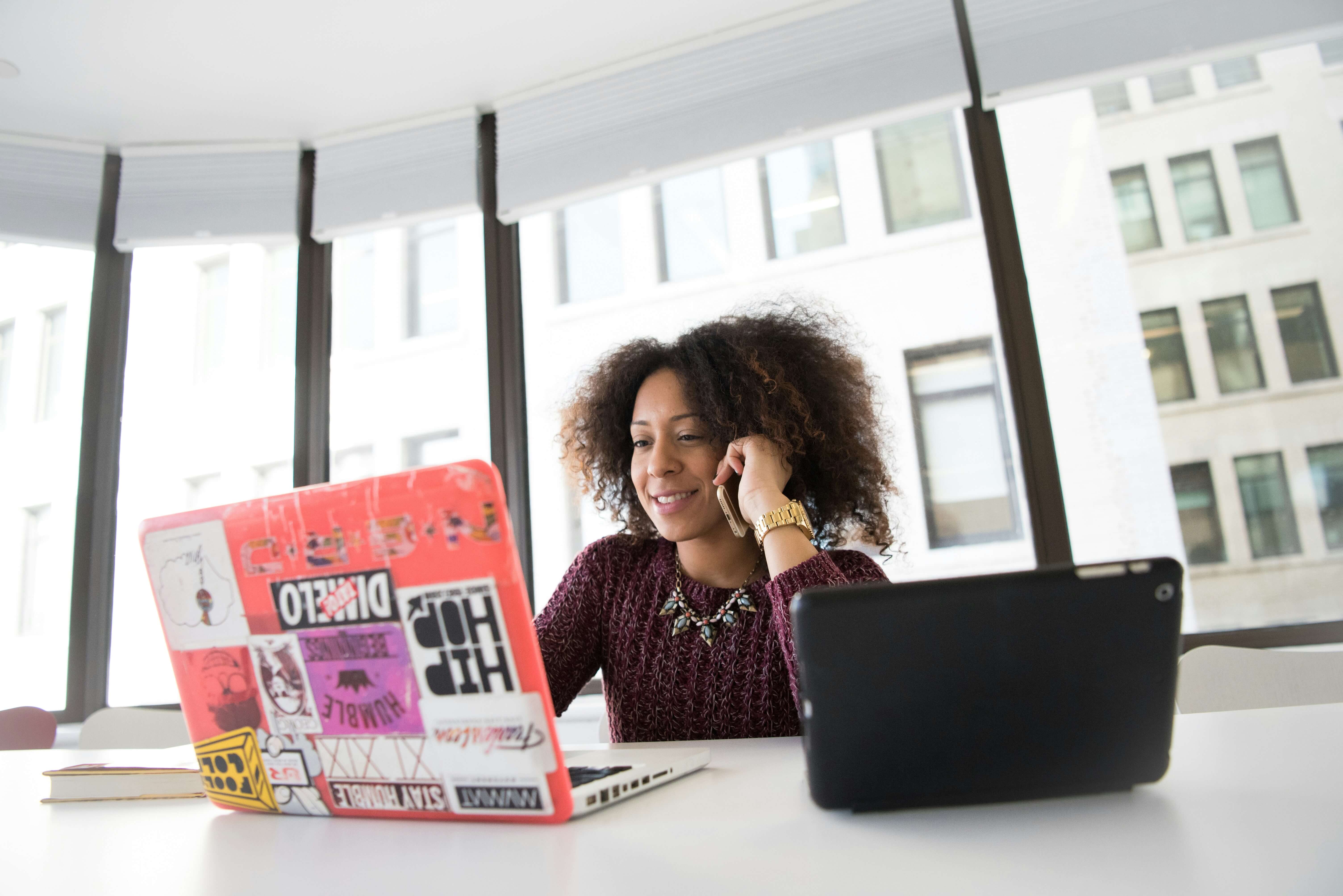 Woman with curly hair on phone, smiling, sits at a table with a laptop covered in stickers and a tablet, in a bright office setting.
