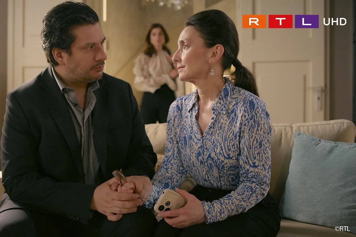 Ein Mann und eine Frau sitzen eng beieinander auf einem Sofa und halten Händchen, während im Hintergrund eine Frau steht.