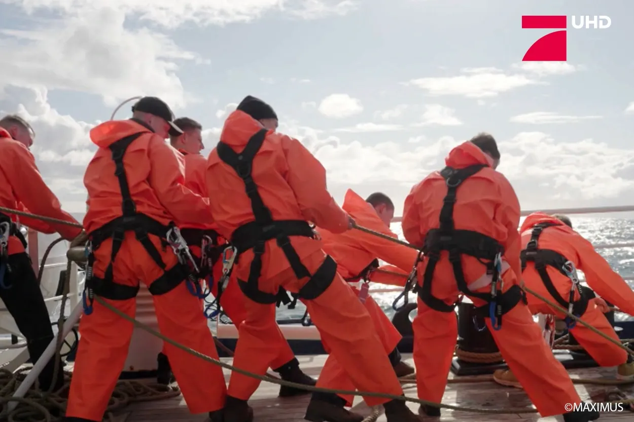 Unter einem bewölkten Himmel ziehen Besatzungsmitglieder in orangefarbenen Anzügen auf einem Boot an Seilen.