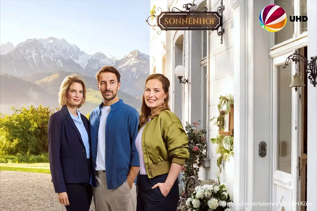 Drei lächelnde Menschen stehen vor einem Gebäude mit der Aufschrift „Sonnenhof“, im Hintergrund sind malerische Berge zu sehen.