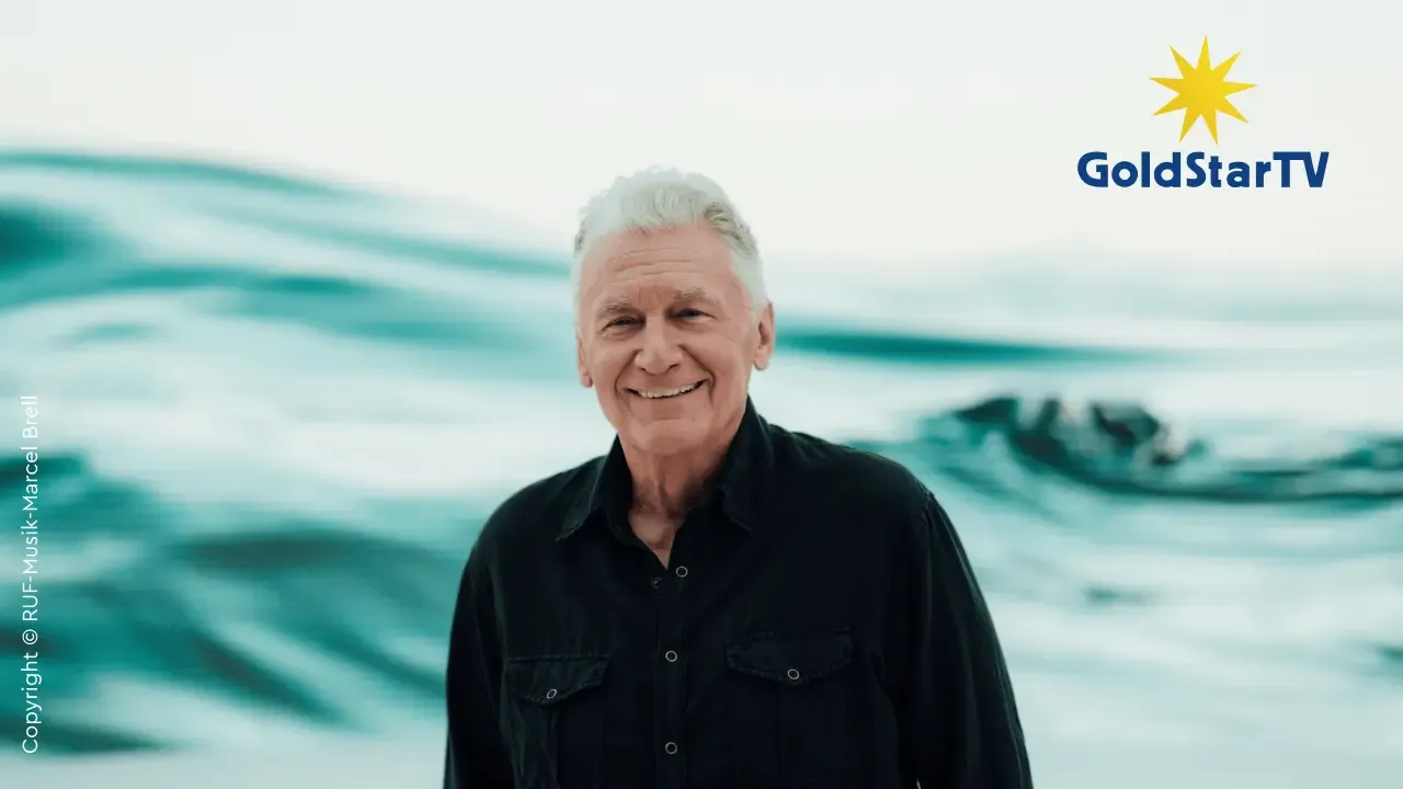 Ein lächelnder Mann mit weißem Haar in einem schwarzen Hemd steht vor einem blauen, abstrakten Hintergrund, auf dem in der oberen rechten Ecke das GoldStarTV-Logo zu sehen ist.
