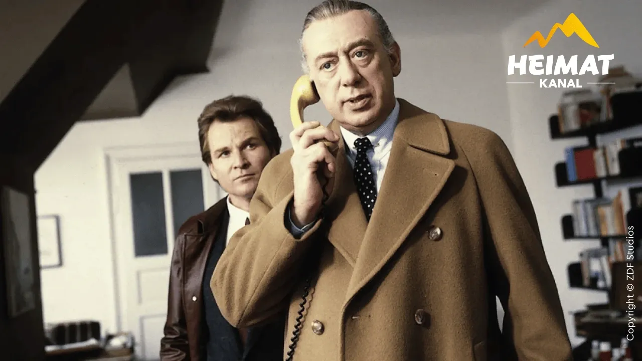 Zwei Männer in Mänteln stehen in einem Büro. Der eine hält einen gelben Telefonhörer in der Hand, während der andere mit aufmerksamem Blick zusieht.
