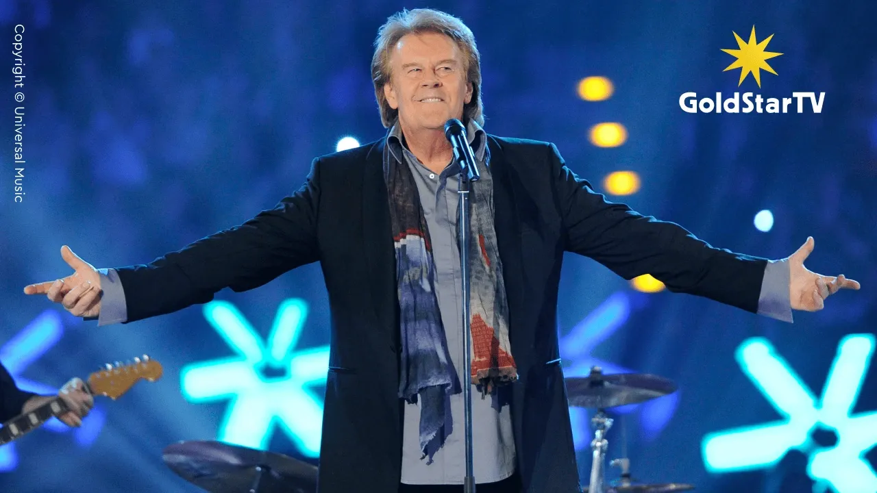 Howard Carpendale singt leidenschaftlich auf der Bühne, die Arme ausgestreckt, mit Schal und Anzug bekleidet, vor einem Mikrofon stehend. Hinter ihm leuchten hellblaue und gelbe Lichter.