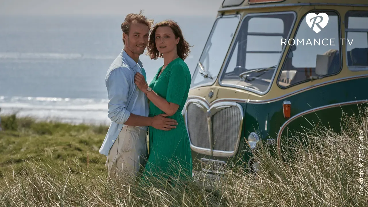 Ein Paar steht sich umarmt neben einem Oldtimer auf einer grasbewachsenen Strandwiese gegenüber, im Hintergrund ist das Meer zu sehen und in der Ecke das Logo von „Romance TV“.