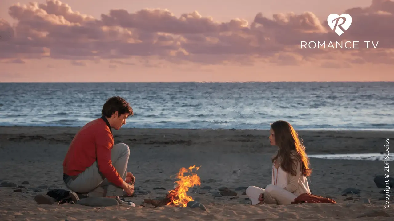 Ein Mann und eine Frau sitzen am Strand neben einem kleinen Lagerfeuer, im Hintergrund sind das Meer und ein rosafarbener Sonnenuntergangshimmel zu sehen.