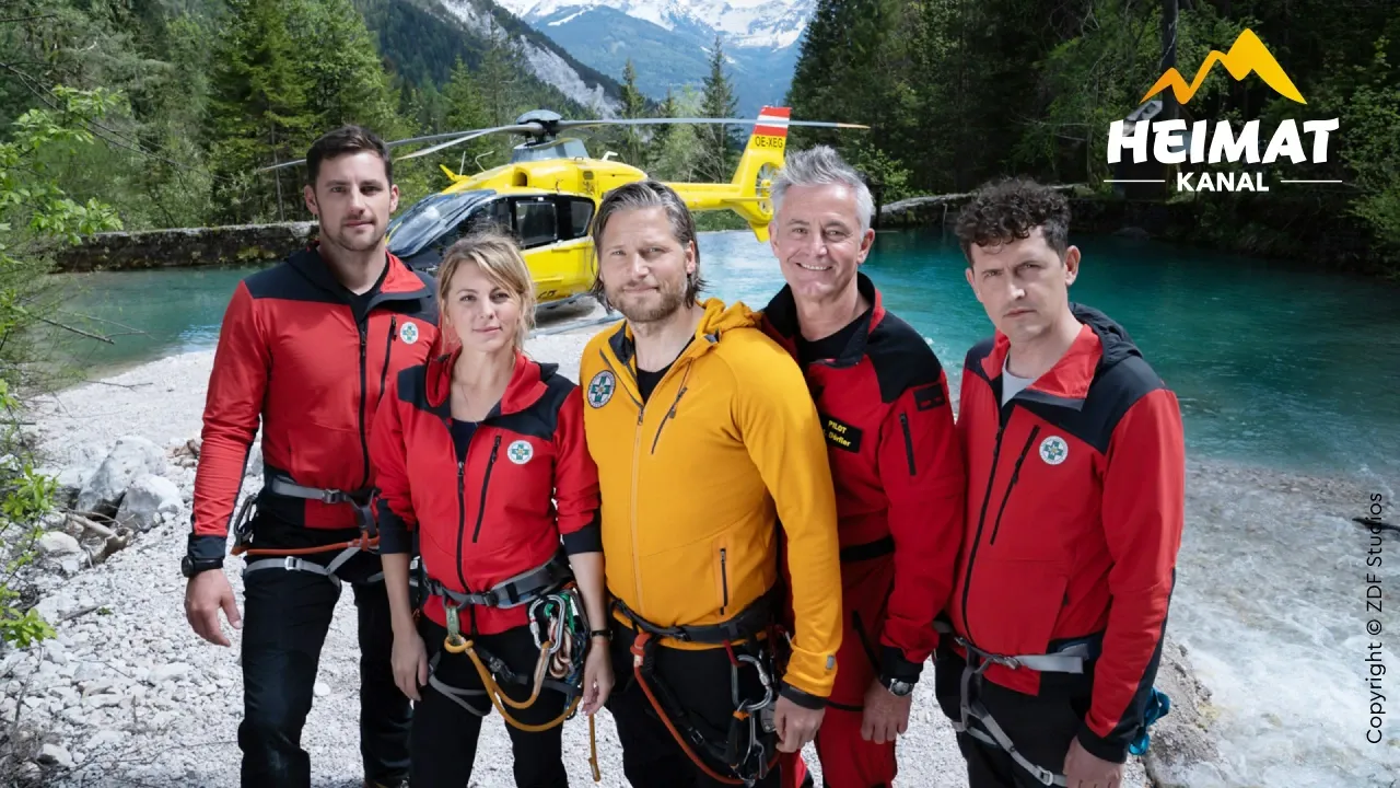 Fünf Mitglieder eines Rettungsteams in bunter Ausrüstung stehen vor einem gelben Hubschrauber an einem Fluss in einer bergigen Waldlandschaft.