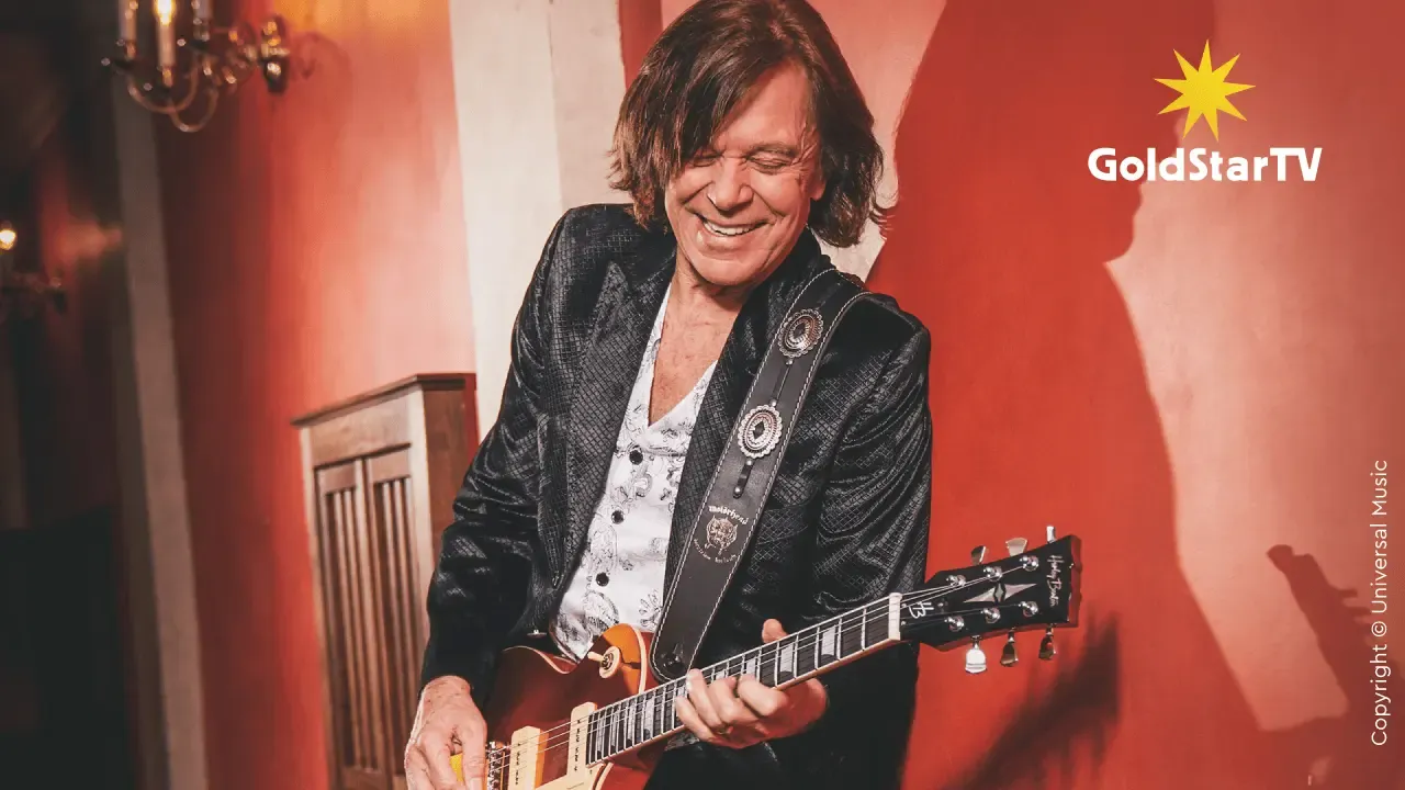 Jürgen Drews in einer dunklen Jacke, der eine E-Gitarre spielt und lächelt. Im Hintergrund eine rote Wand mit dem GoldStarTV-Logo in der rechten oberen Ecke.