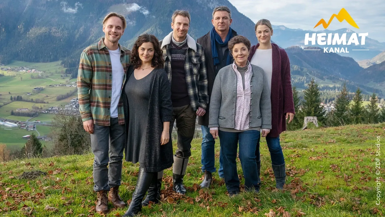 Eine Gruppe von sechs Personen – vier Männer und zwei Frauen – steht auf einem grasbewachsenen Hügel, im Hintergrund erstreckt sich eine malerische Berglandschaft.