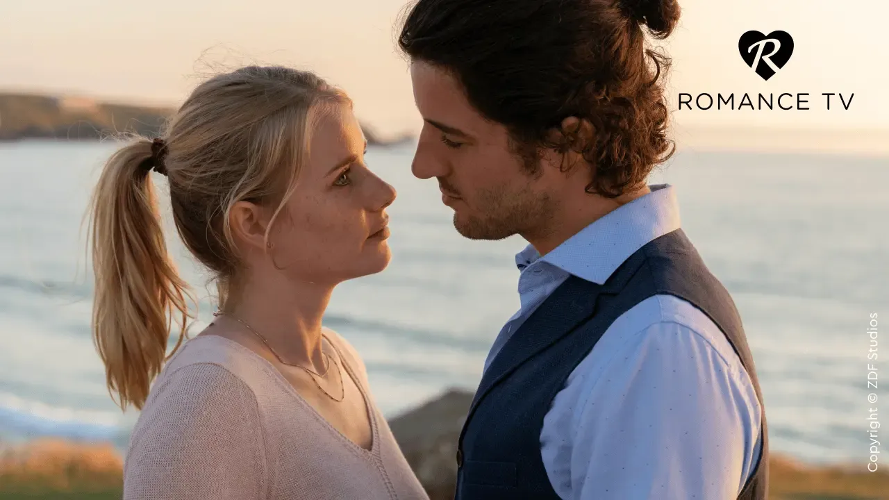 Ein Paar blickt sich bei Sonnenuntergang am Meer in die Augen, während in der Ecke das Logo von „Romance TV“ zu sehen ist.