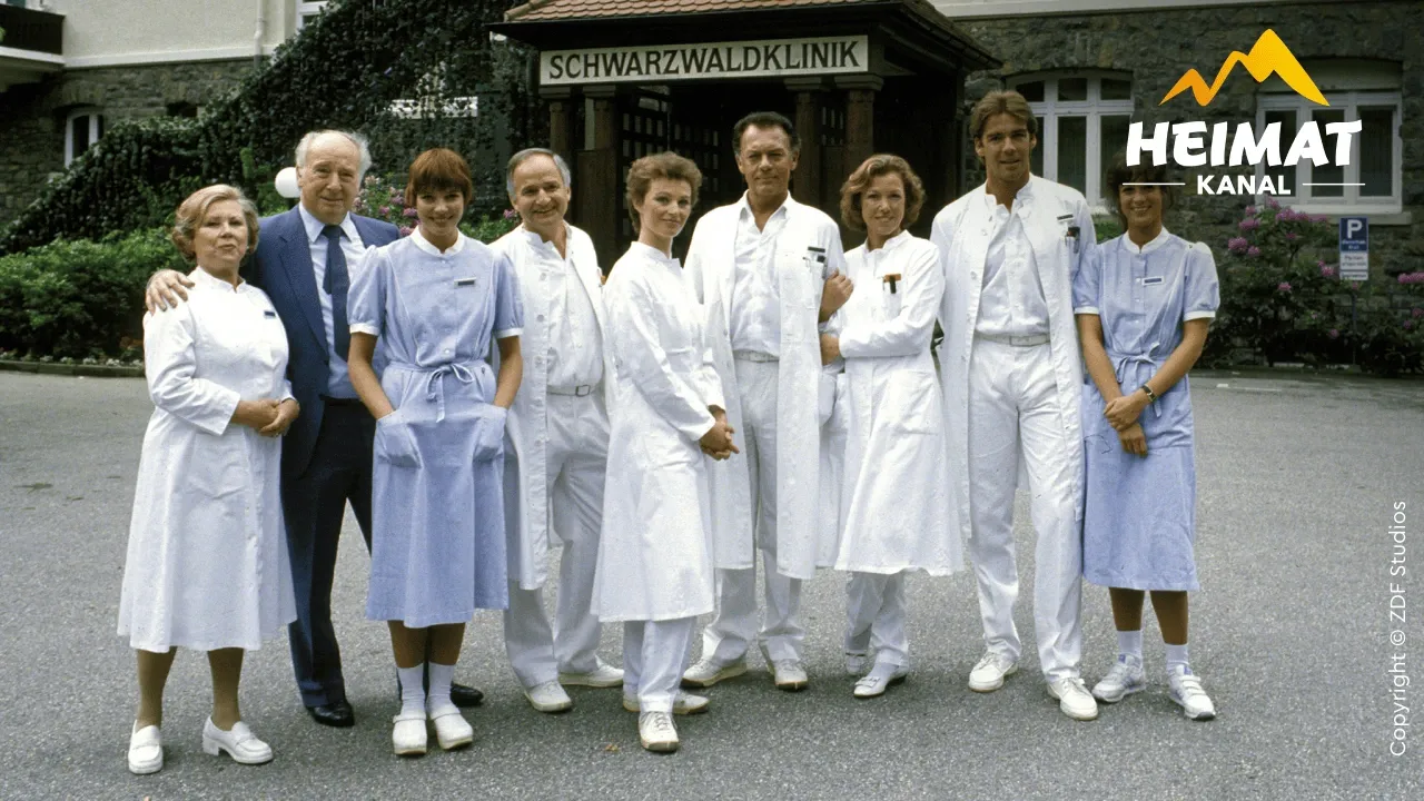 Eine Gruppe von neun Personen, die als Krankenhauspersonal verkleidet sind, steht gemeinsam vor einem Gebäude, an dem ein Schild mit der Aufschrift „Schwarzwaldklinik“ angebracht ist.