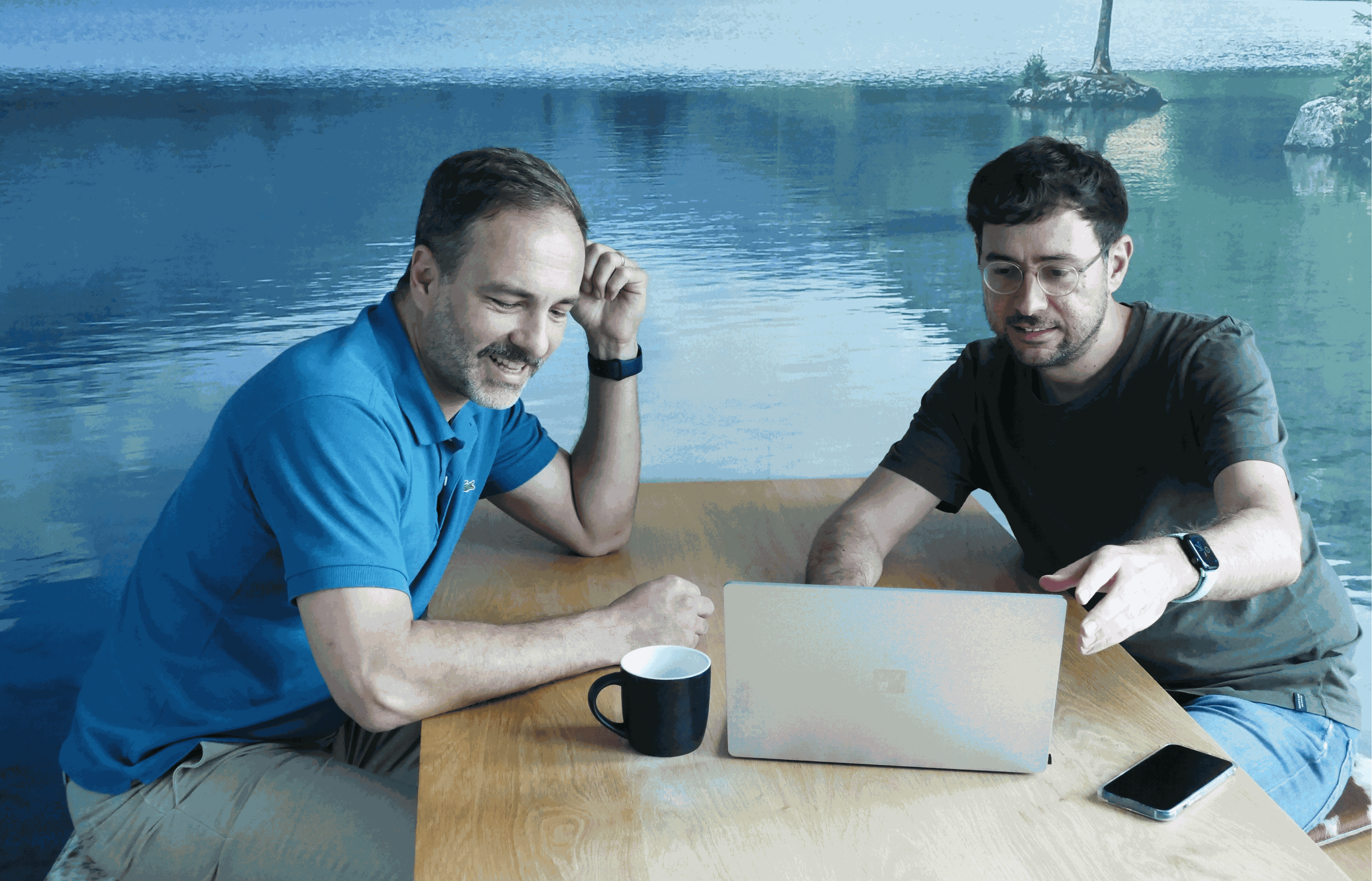 Zwei Männer sitzen an einem Tisch, lächeln und schauen auf einen Laptop. Einer zeigt auf den Bildschirm. Auf dem Tisch stehen eine Tasse und ein Telefon. Im Hintergrund ist ein See zu sehen.