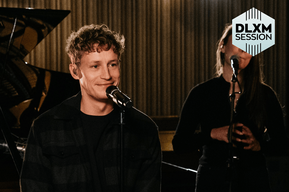 Tim Bendzko singt während einer DLXM-Session in ein Mikrofon, im Hintergrund sind eine Frau und ein Klavier zu sehen.