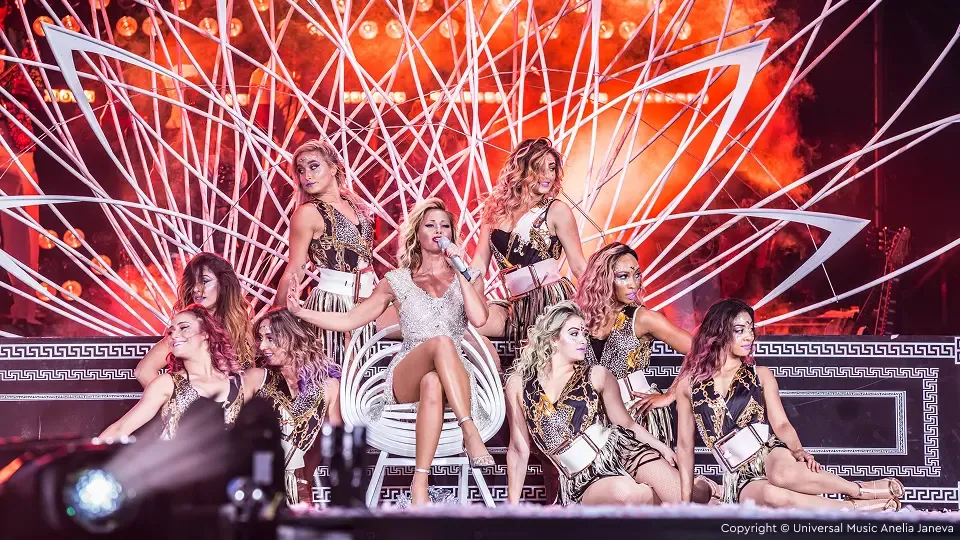 Eine Darstellerin in einem funkelnden Outfit sitzt auf der Bühne, umgeben von Tänzern in Animal-Print, vor einem leuchtenden, farbenfrohen Hintergrund, der an Sonnenstrahlen erinnert.