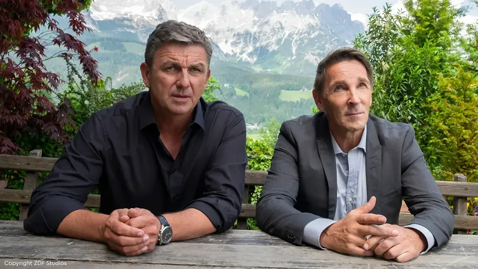 Zwei Männer sitzen draußen an einem Holztisch, der eine in einem schwarzen Hemd, der andere im Anzug, im Hintergrund sind Berge und Grünflächen zu sehen.
