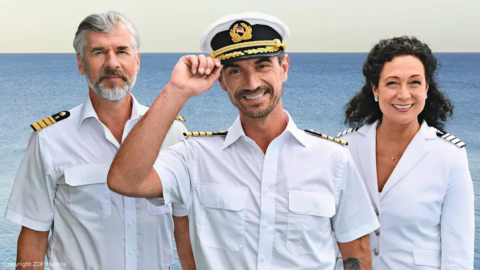 Drei Personen in Marineuniformen lächeln; eine salutiert, vor dem Hintergrund des Meeres.
