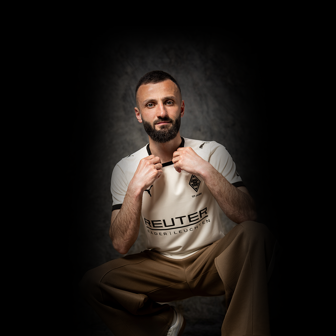 Ein Mann mit Bart hockt vor einem strukturierten grauen Hintergrund und hält den Kragen eines weißen Sporttrikots mit schwarzen Akzenten.