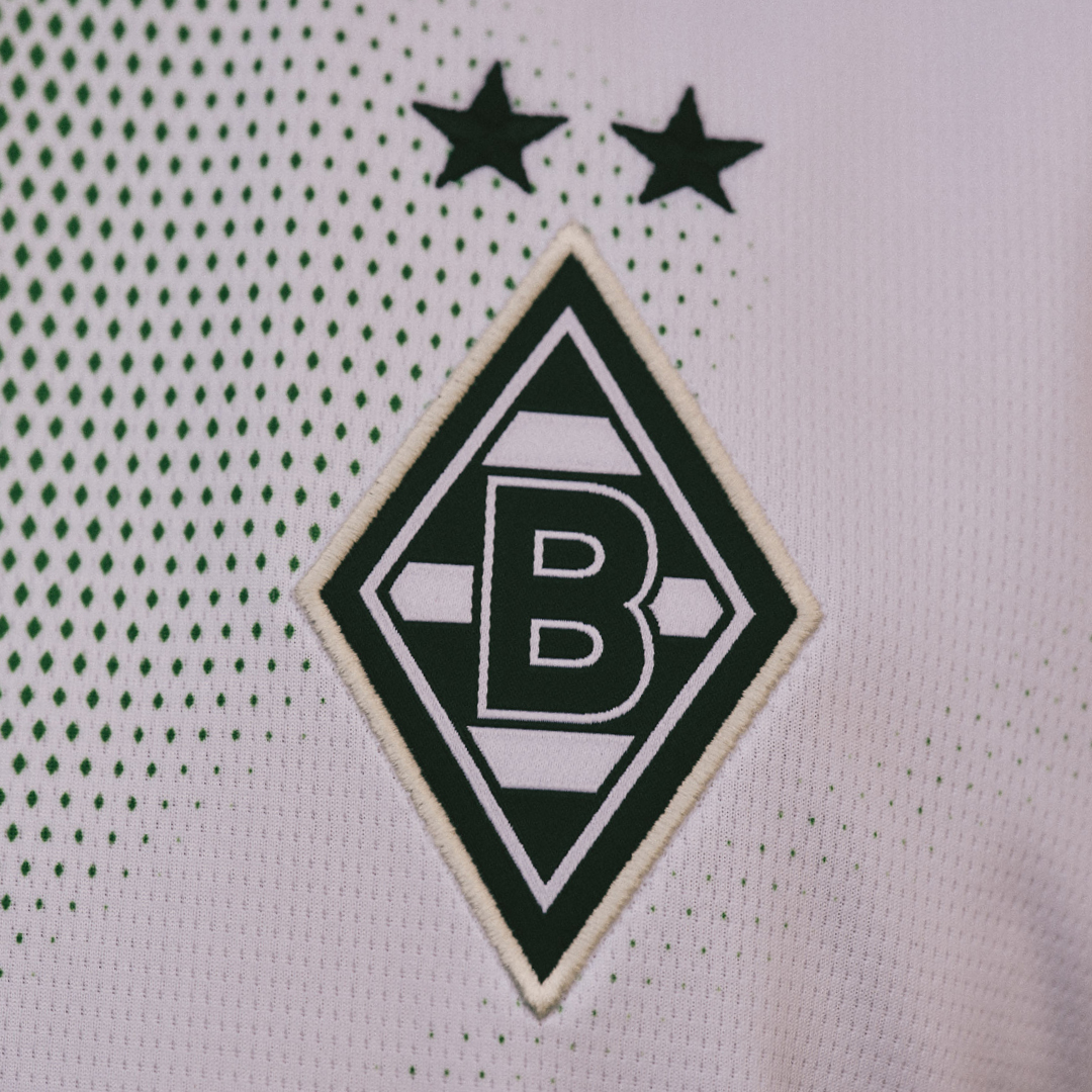 Green and white Borussia Mönchengladbach logo with two stars above on a dotted white fabric background.