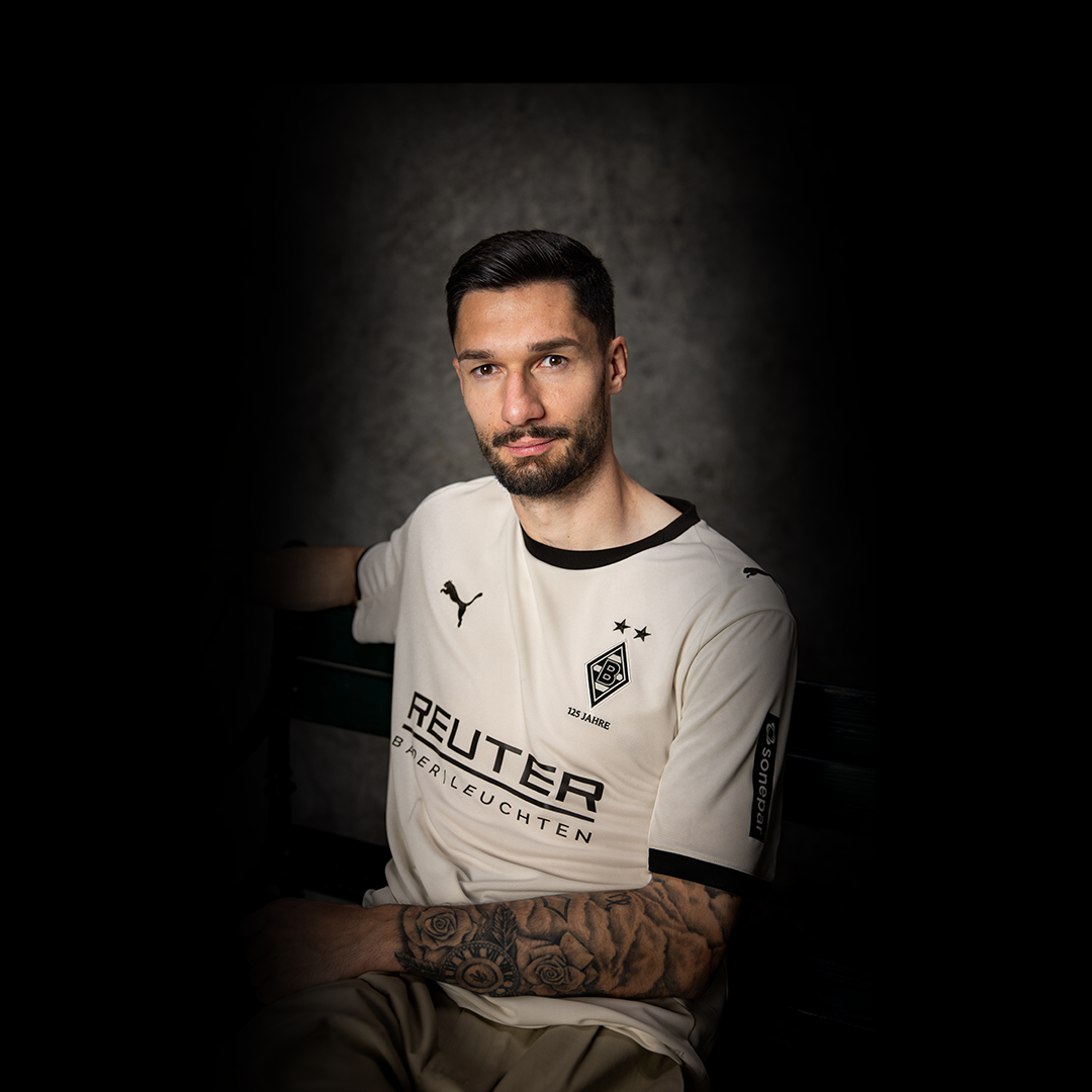 Ein Mann mit Bart sitzt auf einer Bank und trägt ein weißes Fußballtrikot mit Sponsorenlogos vor einem grau strukturierten Hintergrund.